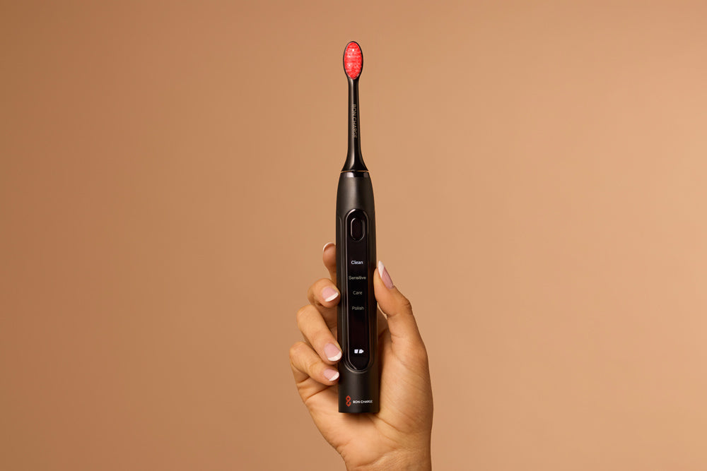 Hand holding a black electric toothbrush with a red bristle head against a brown background